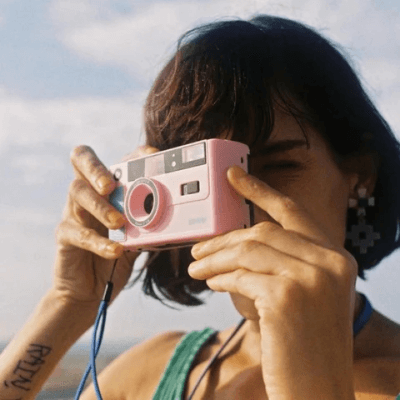 A woman holding a pink film camera up to her eye, capturing a photo outdoors on a sunny day